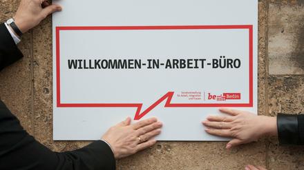 "Willkommen-in-Arbeit-Büro" steht auf dem Schild vor einem Hangar im Flughafen Tempelhof, das zwei Mitarbeiter an eine Wand halten. 