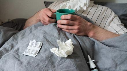 Ein Mann liegt mit Taschentüchern, Teetasse, Nasenspray und Tabletten im Bett. (gestellte Szene)