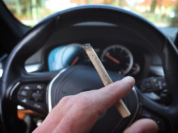 Ein Cannabis-Konsument sitzt mit einem Joint zwischen den Fingern am Steuer eines Autos. 