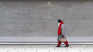 Eine Frau mit Maske geht an einem mit Rolltor verschlossenen Geschäft vorbei.