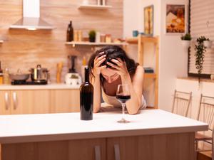 Eine Frau sitzt am Küchentisch und stützt ihren Kopf mit beiden Händen auf der Tischplatte ab. Vor ihr stehen eine Weinflasche und ein halbvolles Weinglas. 