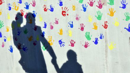 Auf einer Betonwand mit bunten Abdrücken von Kinderhänden ist ein Mann mit Kind auf dem Arm und an der Hand zu sehen.