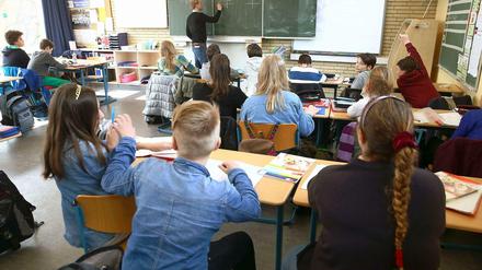 Schülerinnen und Schüler sitzen in einem Klassenraum, ein Lehrer schreibt etwas an die Tafel.